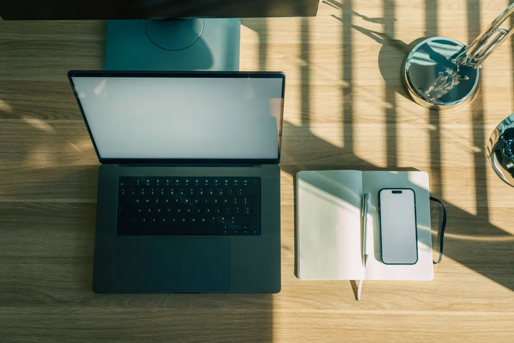 A laptop and a phone on a sunlit work desk